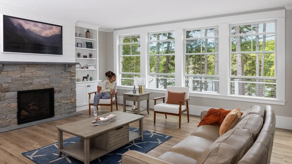 Interior living room, showing a woman looking outside with a forest view. With open white framed double-hung windows.